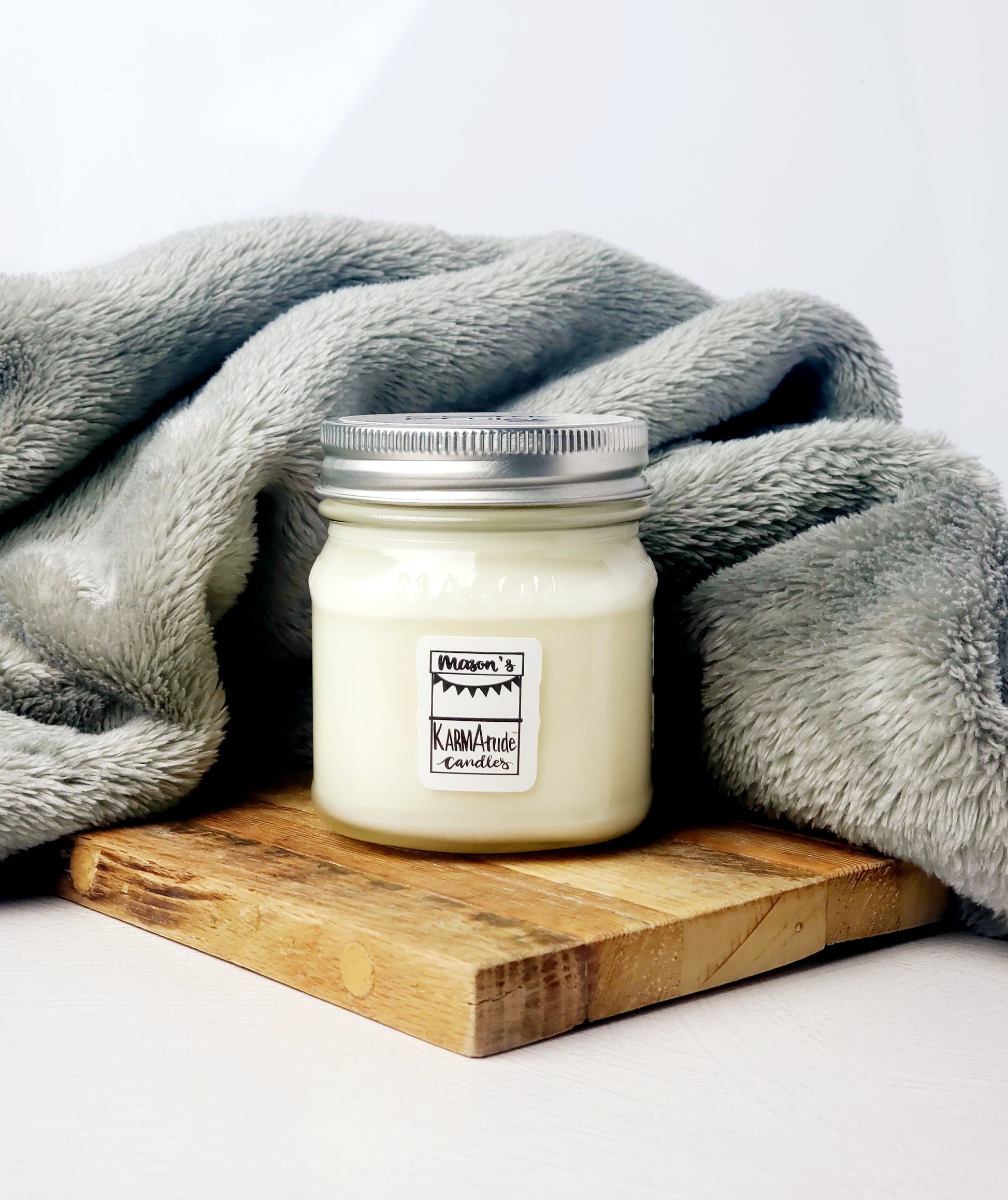 Mason jar glass Karmatude Candle against a gray blanket on a wood trivet.