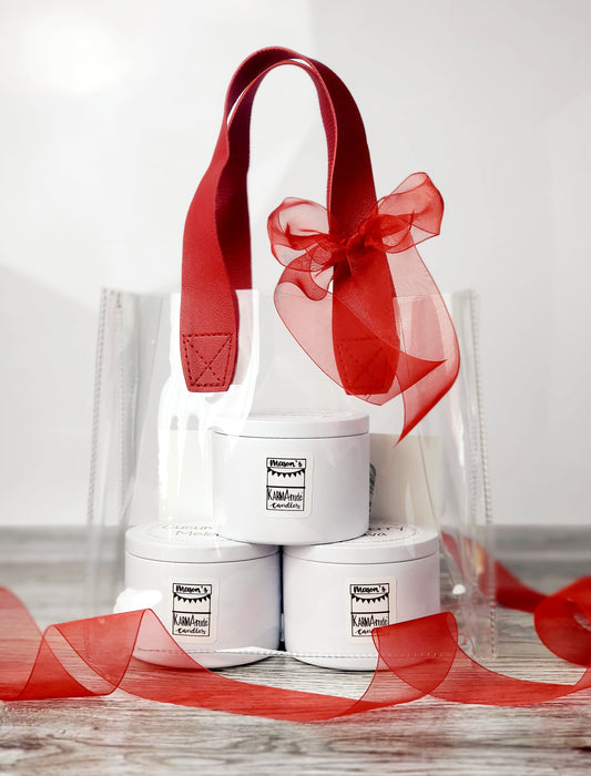 Clear vinyl bag with red handles and red bow containing three 8oz white tin Karmatude Candles.