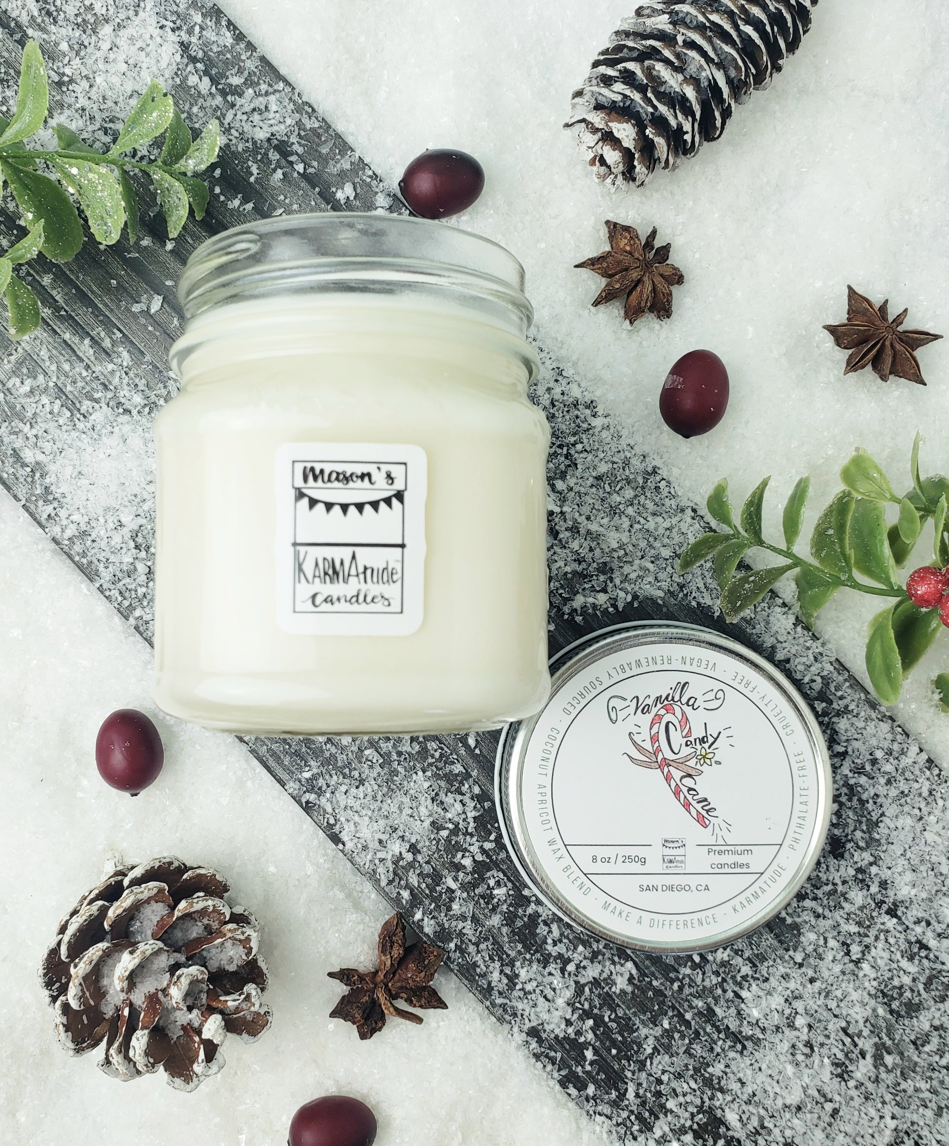 Picture of clear glass Mason Jar candle in Vanilla Candy Cane with metal lid on wood and snow platform surrounded by pine cones, star anise, cranberries, and red berries. 