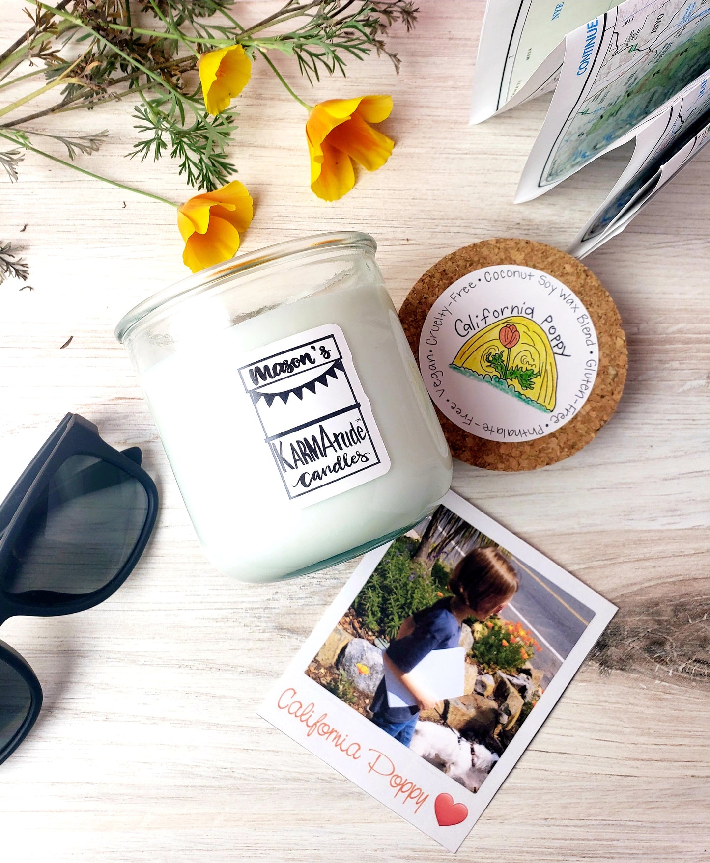 Photo of California Poppy Karmatude candle in clear glass jar with cork lid surrounded by golden poppies, map, sunglasses, and photos of boy walking a dog.