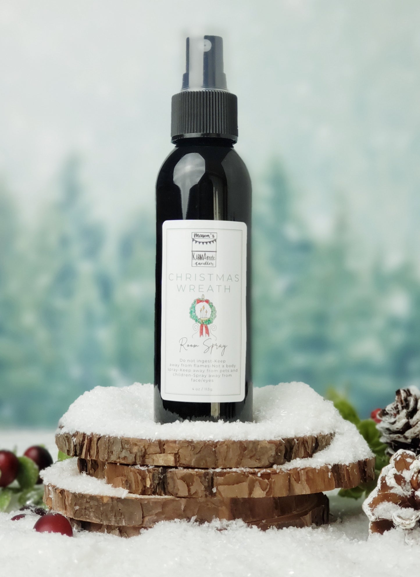 Christmas Wreath room spray in black bottle with lid on stacked wood coasters in the snow with berries and snow cones.