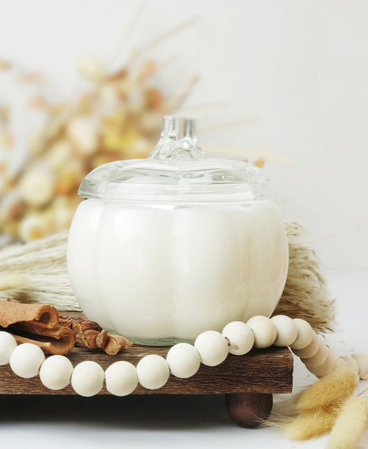 Clear Glass pumpkin candle with lid surrounded by wood beads, cinnamon, and fluffy decor on a wood tray.