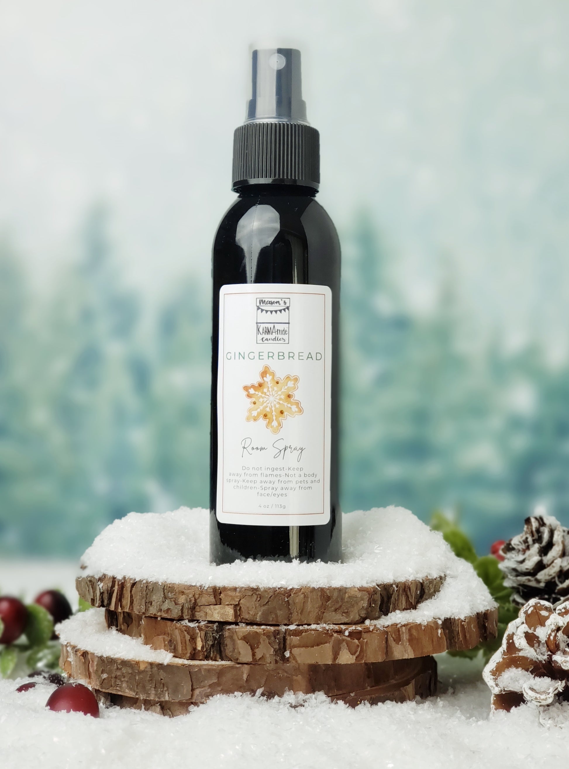 Gingerbread room spray in black bottle with lid on stacked wood coasters in the snow with berries and snow cones.