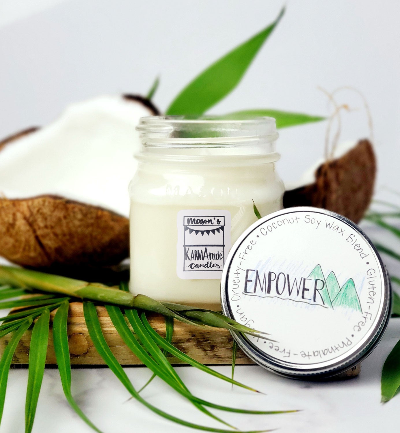 Karmatude Candles Empower candle in clear Mason jar with lid surrounded by palm fronds and coconut.