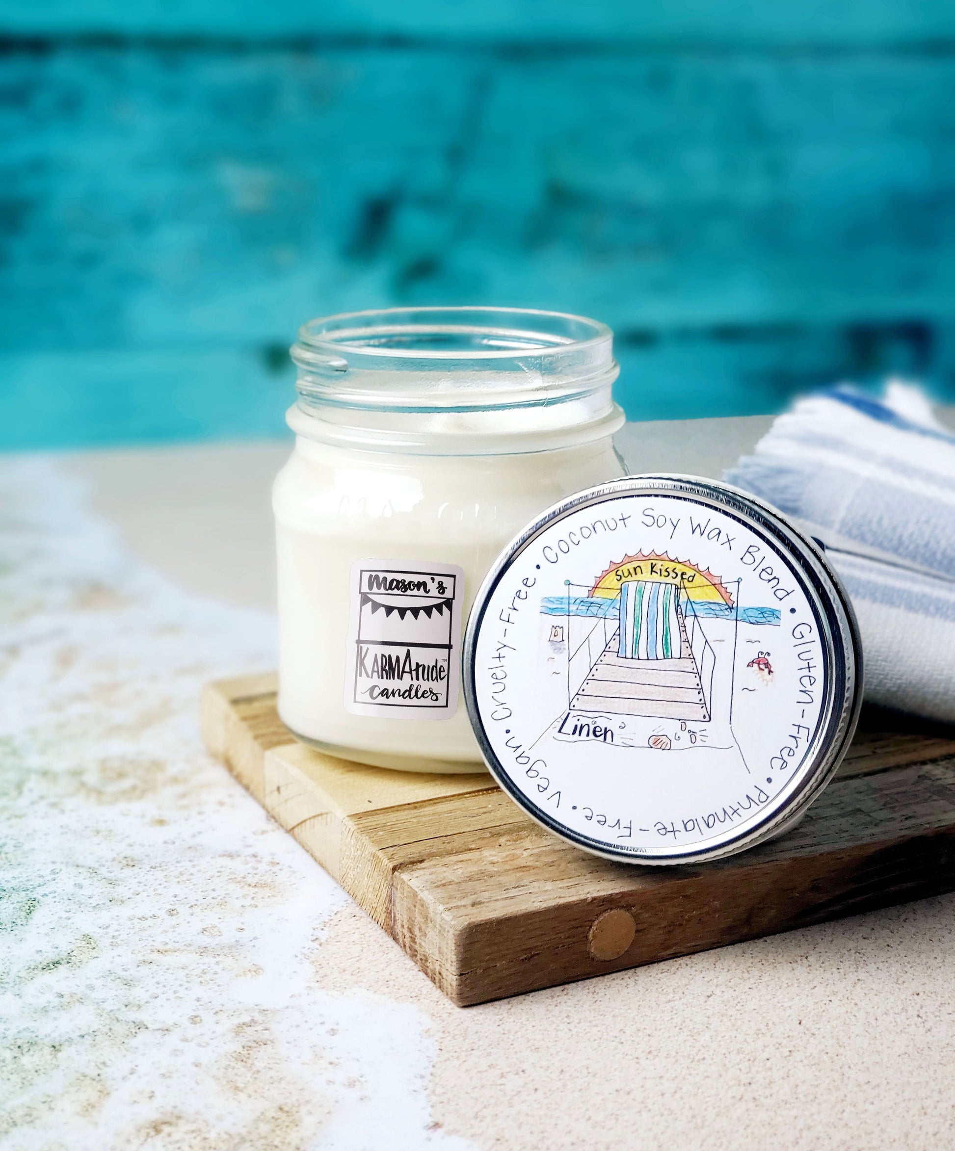 Sun Kissed Linen  Karmatude Candle in a clear Mason Jar candle with lid on a wood trivet surrounded by beach towel, sand and water.