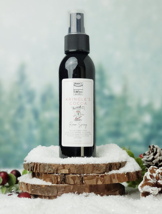 Kringle's Cocoa room spray in black bottle with lid on stacked wood coasters in the snow with berries and snow cones.