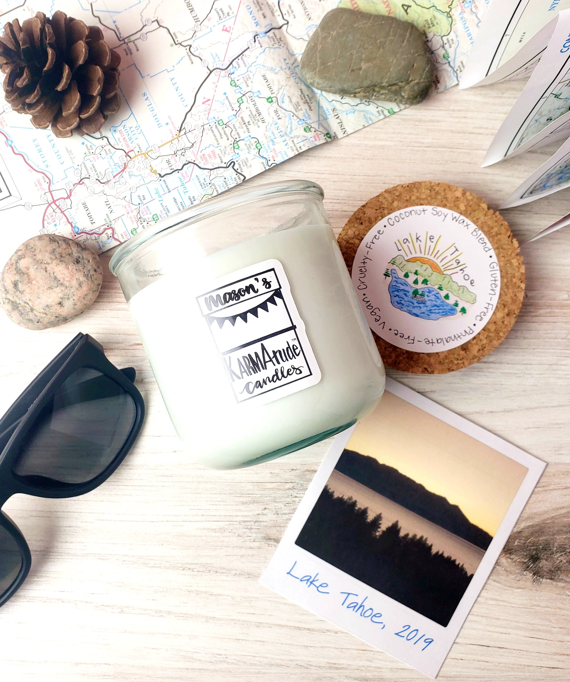 Photo of Lake Tahoe  Karmatude candle in clear glass jar with cork lid surrounded by lake, mountains,  rocks, sunglasses, pinecone and map.