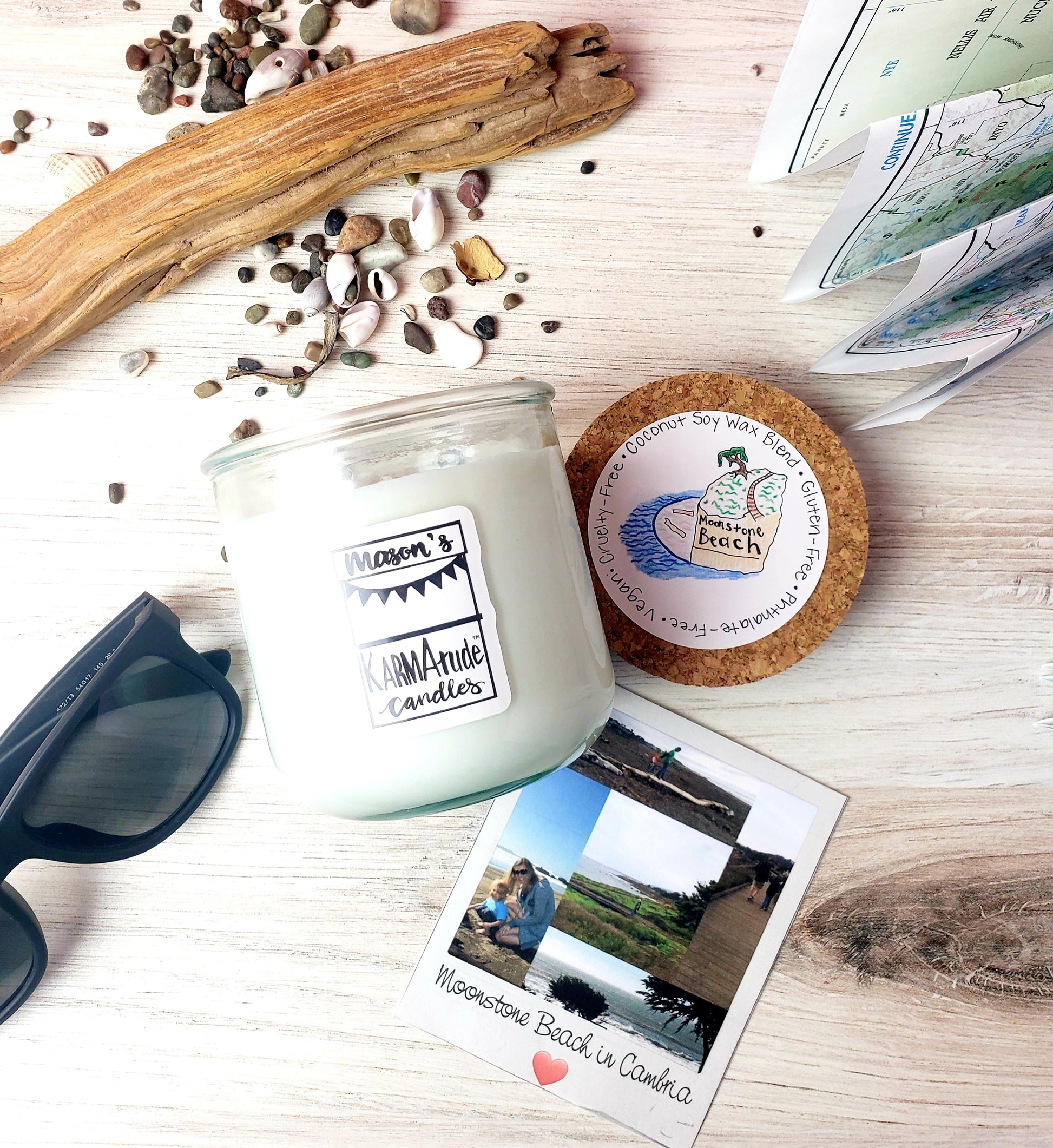 Photo of Moonstone Beach Karmatude candle in clear glass jar with cork lid surrounded by driftwood, small rocks, shells, sunglasses, map and photo of the beach.