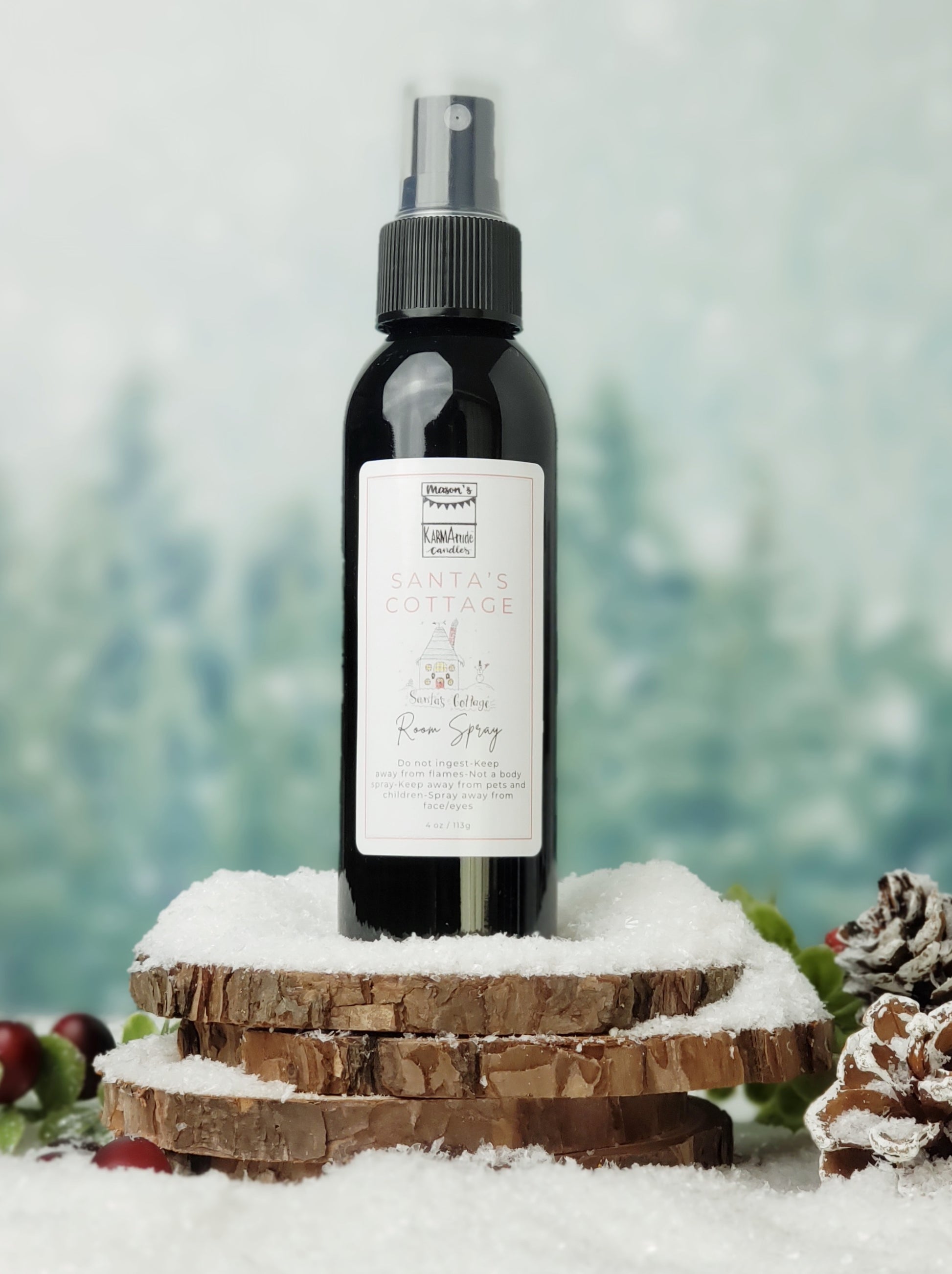 Santa's Cottage room spray in black bottle with lid on stacked wood coasters in the snow with berries and snow cones.