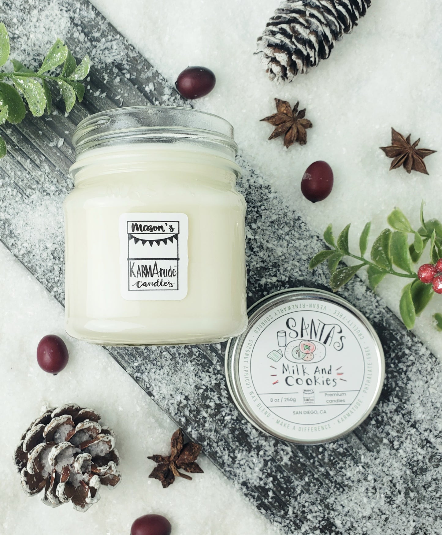 Picture of clear glass Mason Jar candle in Santa's Milk and Cookies with metal lid on wood and snow platform surrounded by pine cones, star anise, cranberries, and red berries. 