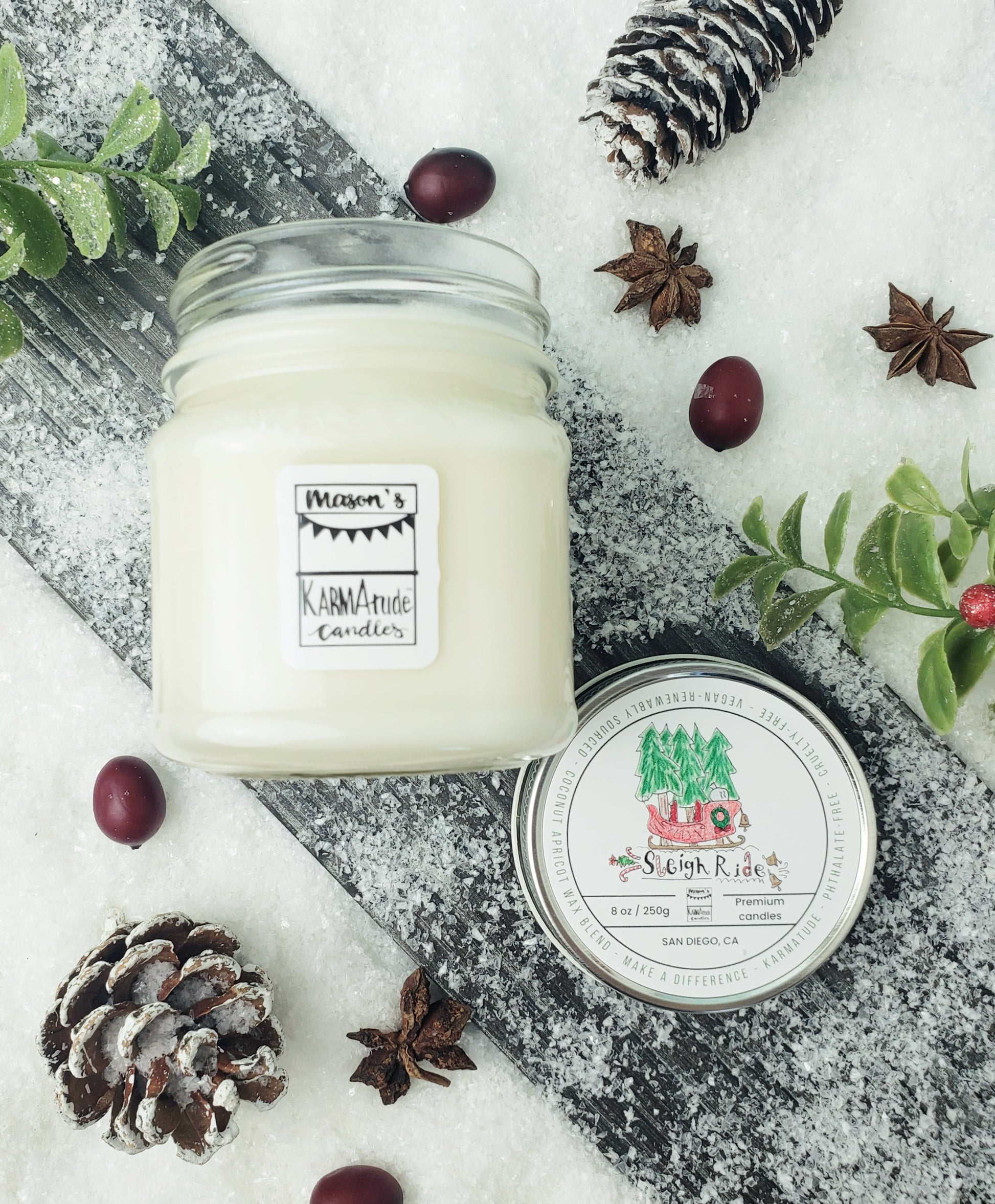 Picture of clear glass Mason Jar candle in Sleigh Ride with metal lid on wood and snow platform surrounded by pine cones, star anise, cranberries, and red berries. 
