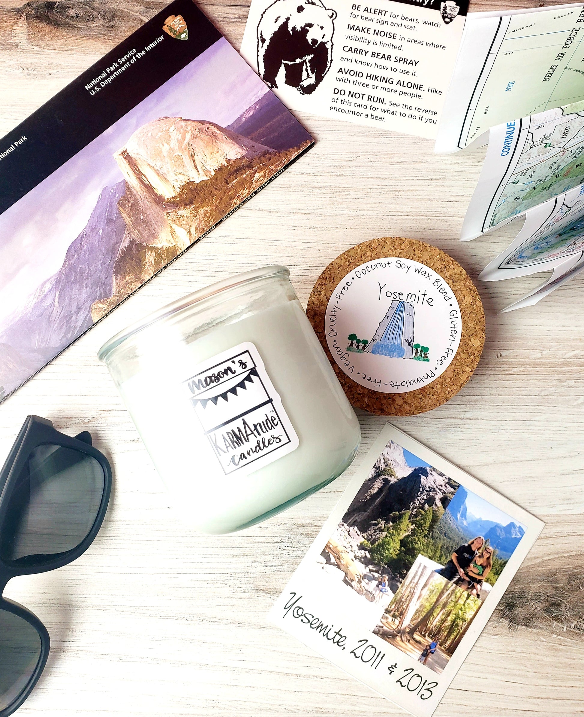 Photo of Yosemite Karmatude candle in clear glass jar with cork lid surrounded by maps, bear warning, sunglasses, and photo of Yosemite with family.